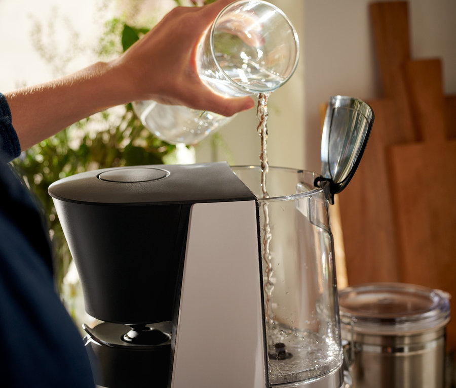 Kobieta nalewa wodę z przezroczystej szklanki do szaro-czarnego ekspresu przelewowego »Let's Brew«.