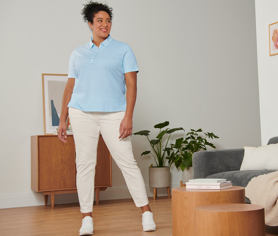 Kobieta pozuje w kremowych spodniach dżinsowych o kroju slim fit, jasnoniebieskiej koszulce polo i białych butach typu sneakers ze skóry welurowej.