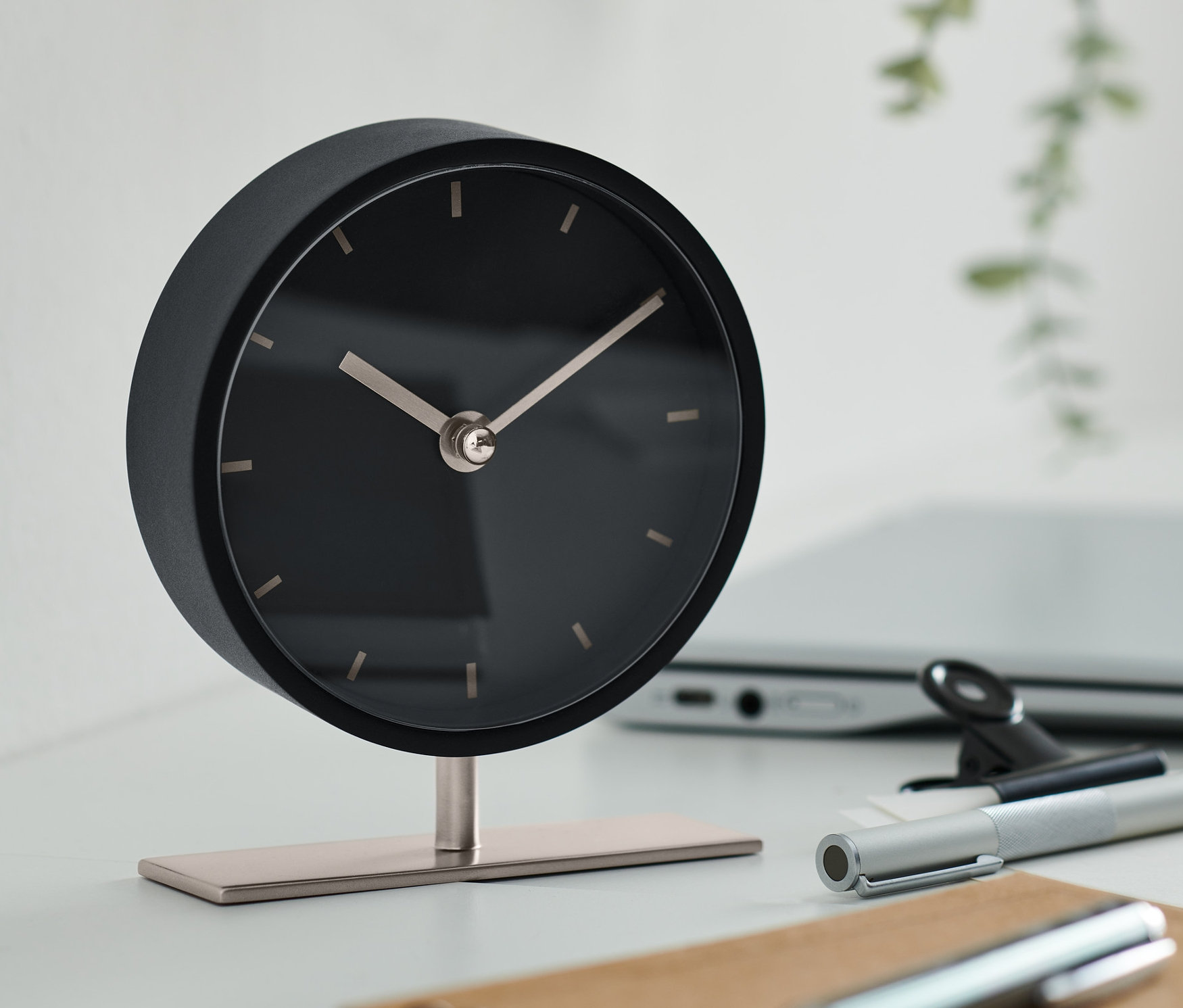 Czarny designerski zegar stołowy stoi na stole z laptopem, długopisem i notatnikiem.