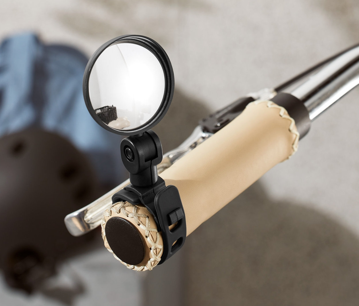 Zbliżenie na kierownicę roweru z zamontowanym małym lusterkiem rowerowym. W tle widać kask i kurtkę.