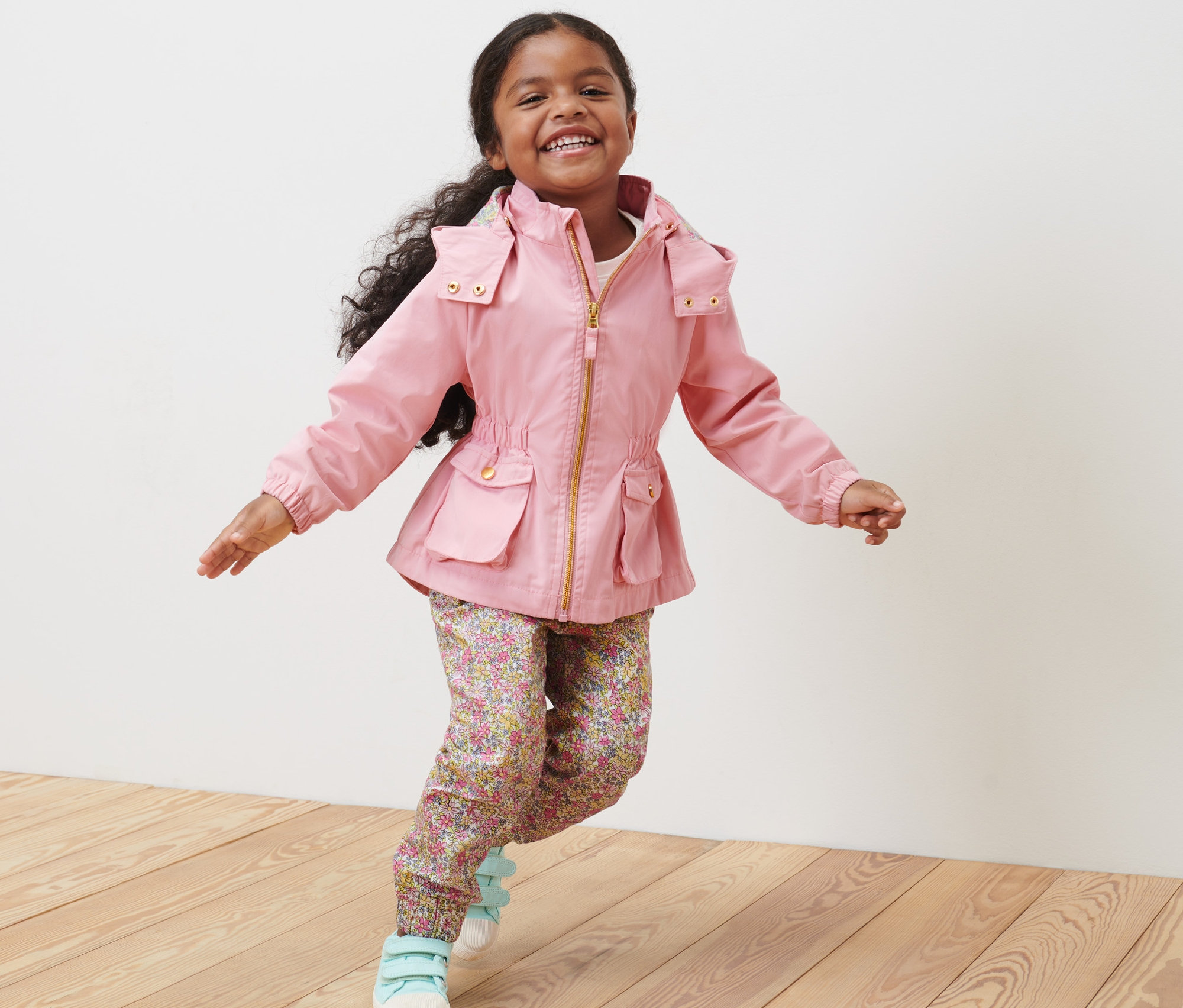 Uśmiechnięta dziewczynka w kurtce typu parka biegnie po drewnianej podłodze.