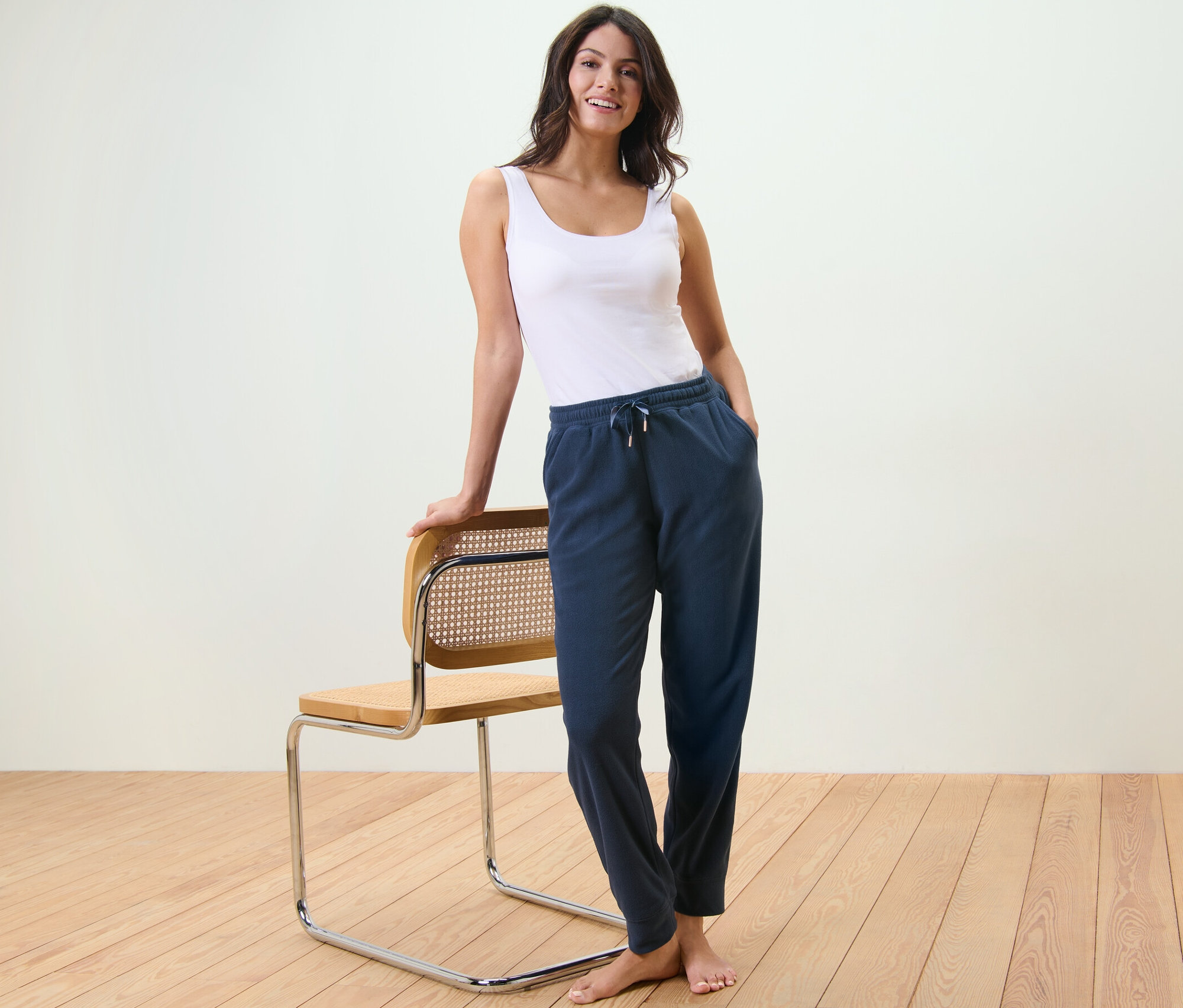 Kobieta stoi oparta o krzesło, ubrana w podkoszulek i spodnie rekreacyjne z polaru.