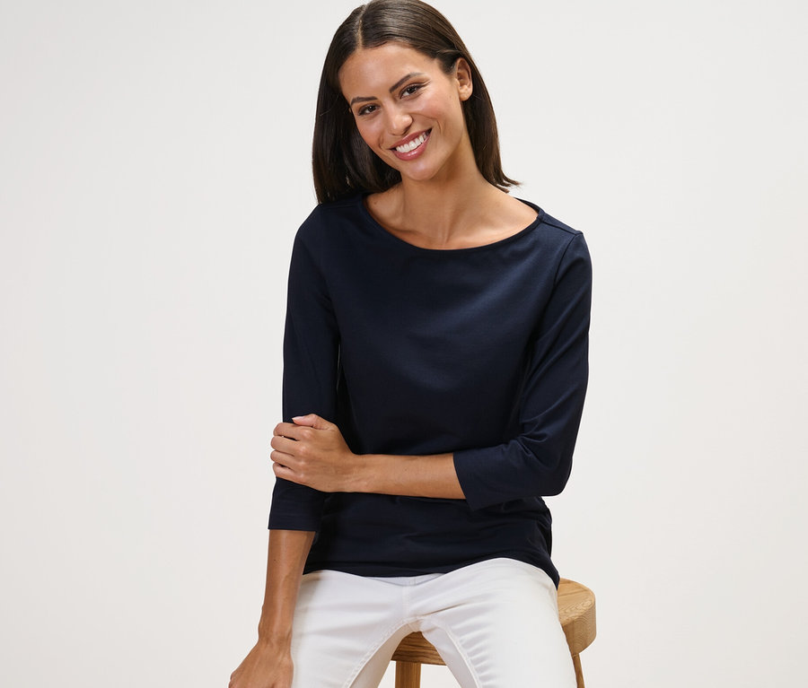 Kobieta siedzi na stołku, ubrana w granatową bluzkę z rękawami o długości 3/4.