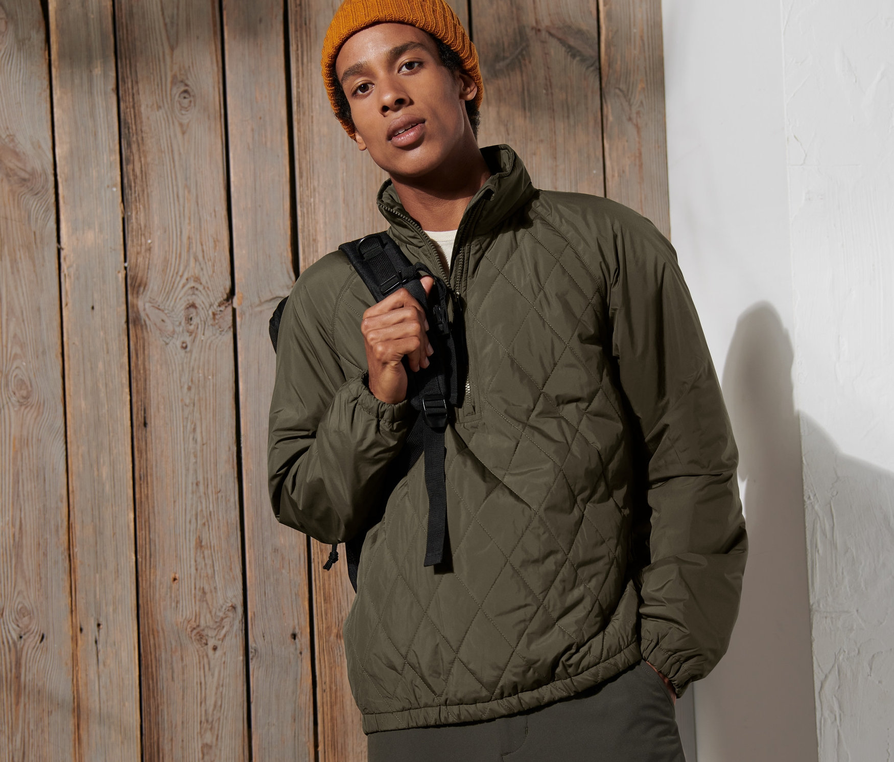 Mężczyzna pozuje na tle drewnianej ściany, ubrany w kurtkę wiatroszczelną i żółtą czapkę.