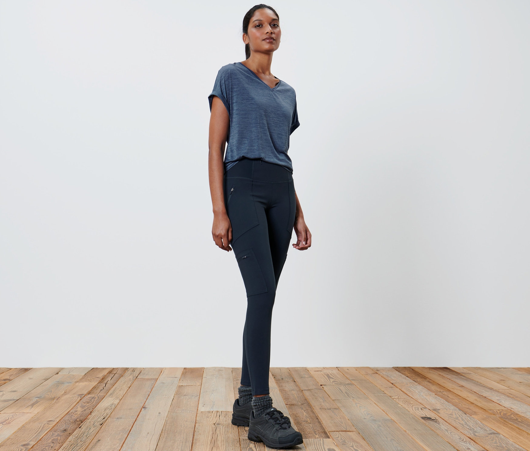 Kobieta w szarej koszulce i czarnych legginsach outdoorowych stoi na drewnianej podłodze.
