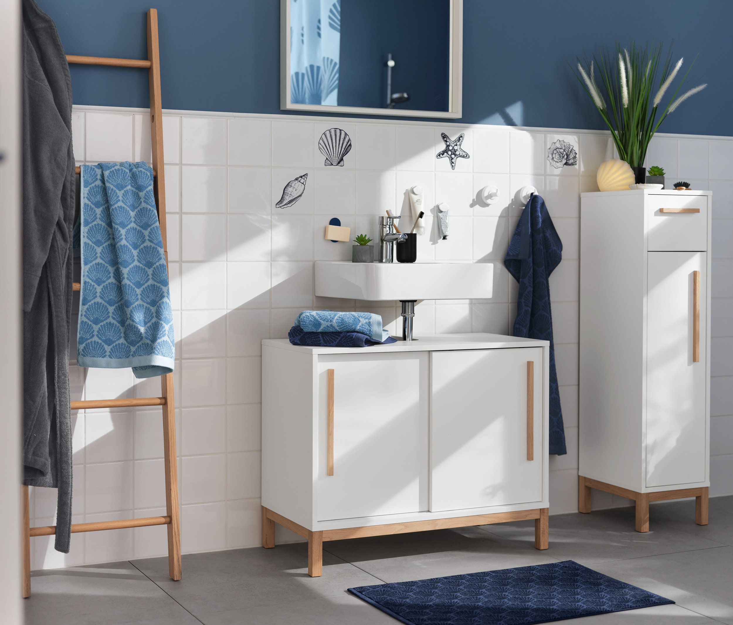 Łazienka z umywalką, lustrem i miejscem do przechowywania. Morskie naklejki na kafelkach zdobią ścianę. Na drewnianej drabinie wisi niebieski ręcznik „Muszla” i szary szlafrok.