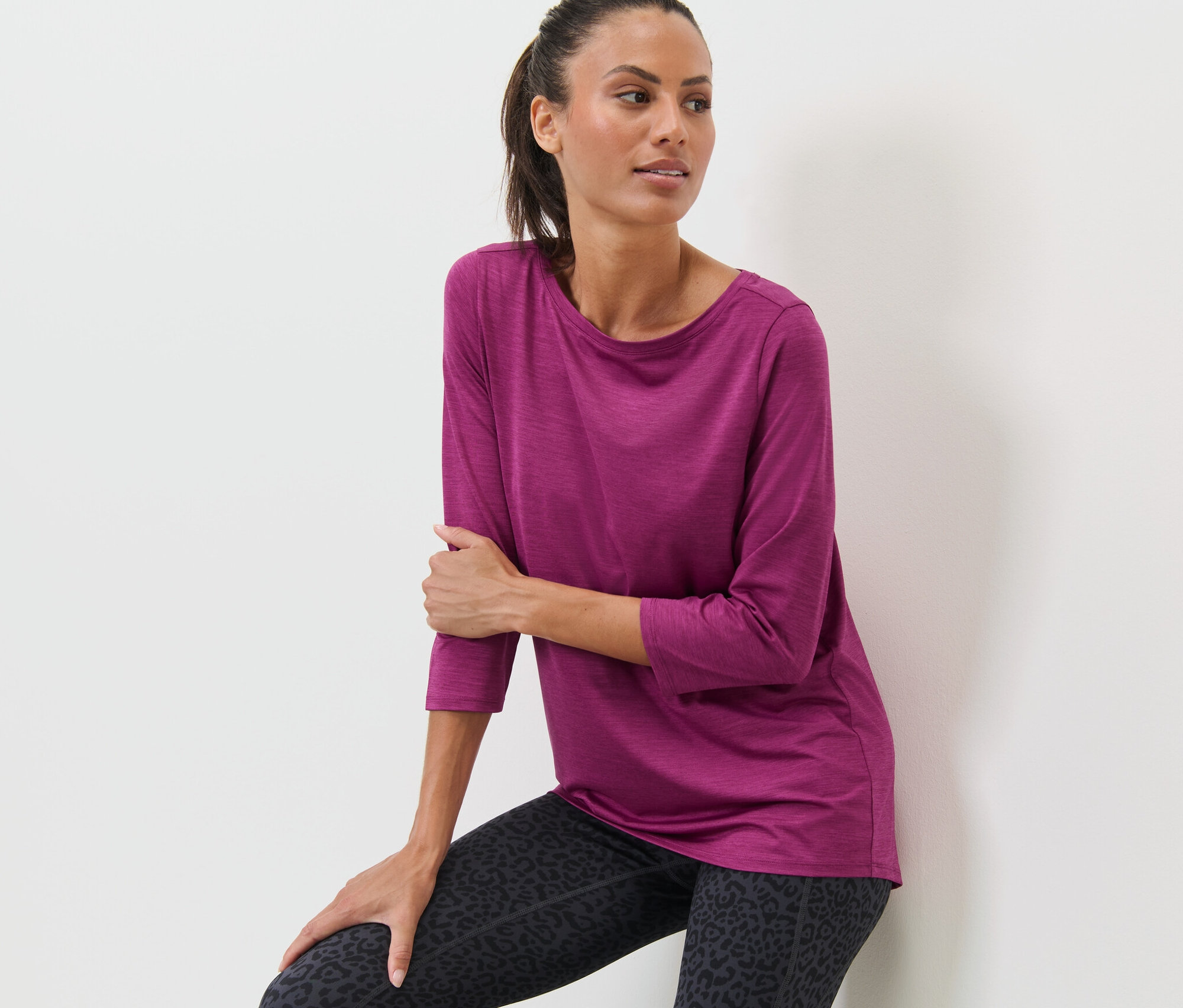 Kobieta oparta o ścianę ma na sobie jagodową koszulkę funkcyjną z rękawem 3/4 i legginsy we wzór zwierzęcy.