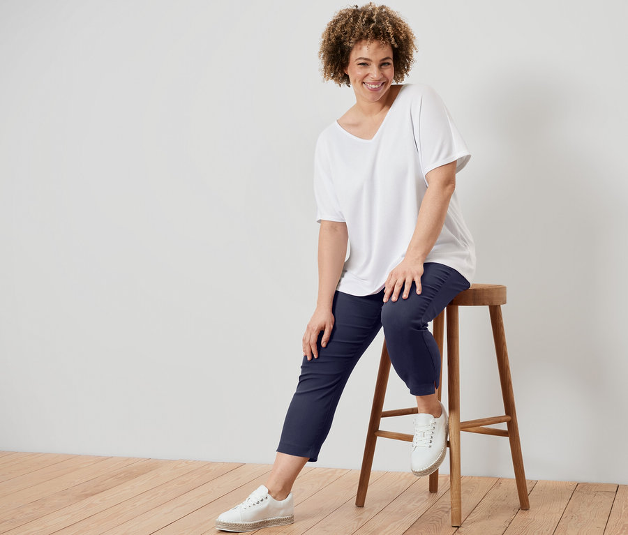 Kobieta siedzi na stołku, ubrana w białą koszulkę z fakturowaną powierzchnią i granatowe spodnie stretchowe o długości 7/8.
