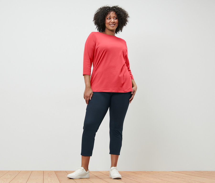 Kobieta pozuje w koralowej bluzce z rękawami 3/4 i granatowych spodniach.