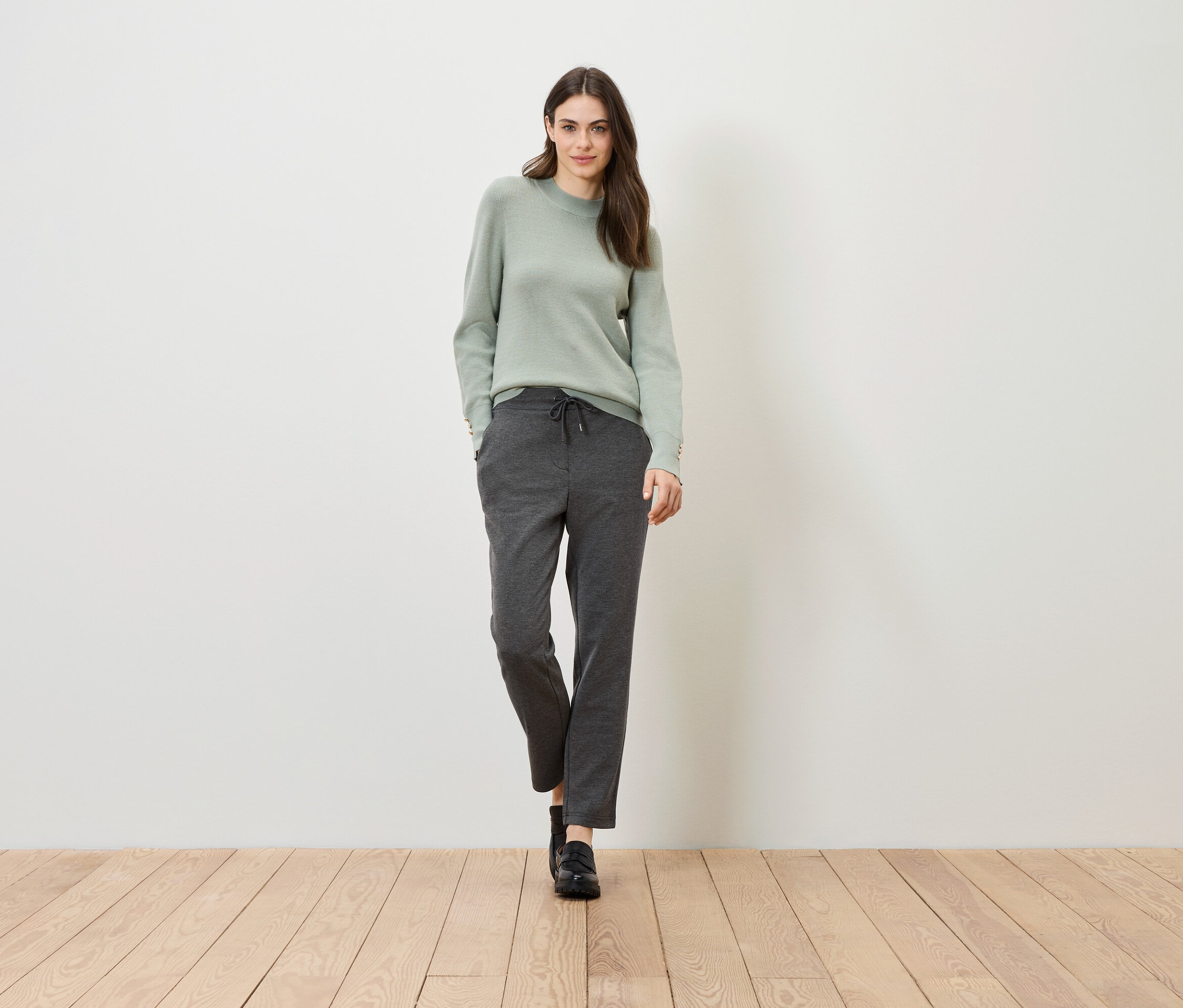 Kobieta stoi na drewnianej podłodze na tle białej ściany. Ma na sobie zielony sweter i szare spodnie.