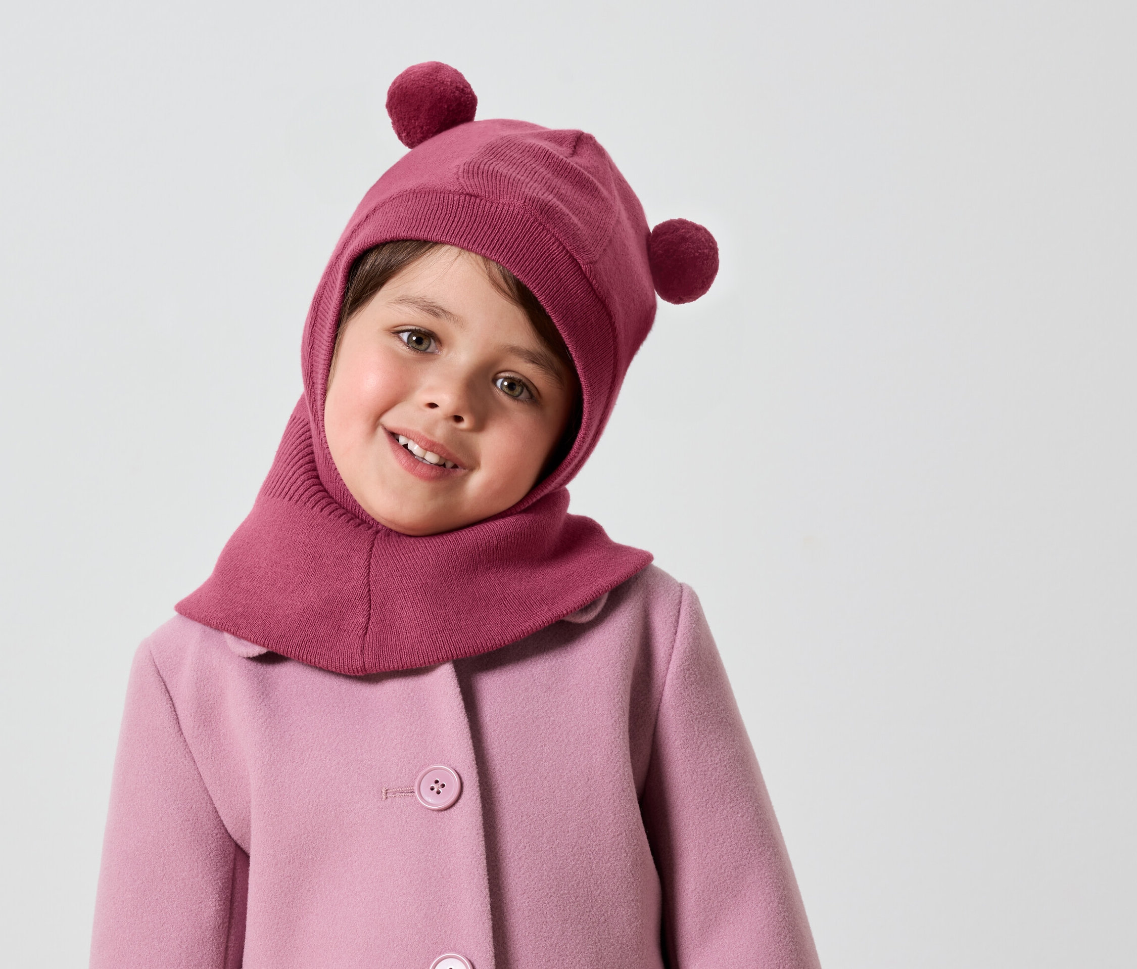 Uśmiechnięta dziewczynka w różowej kominiarce z pomponami i różowym płaszczu.