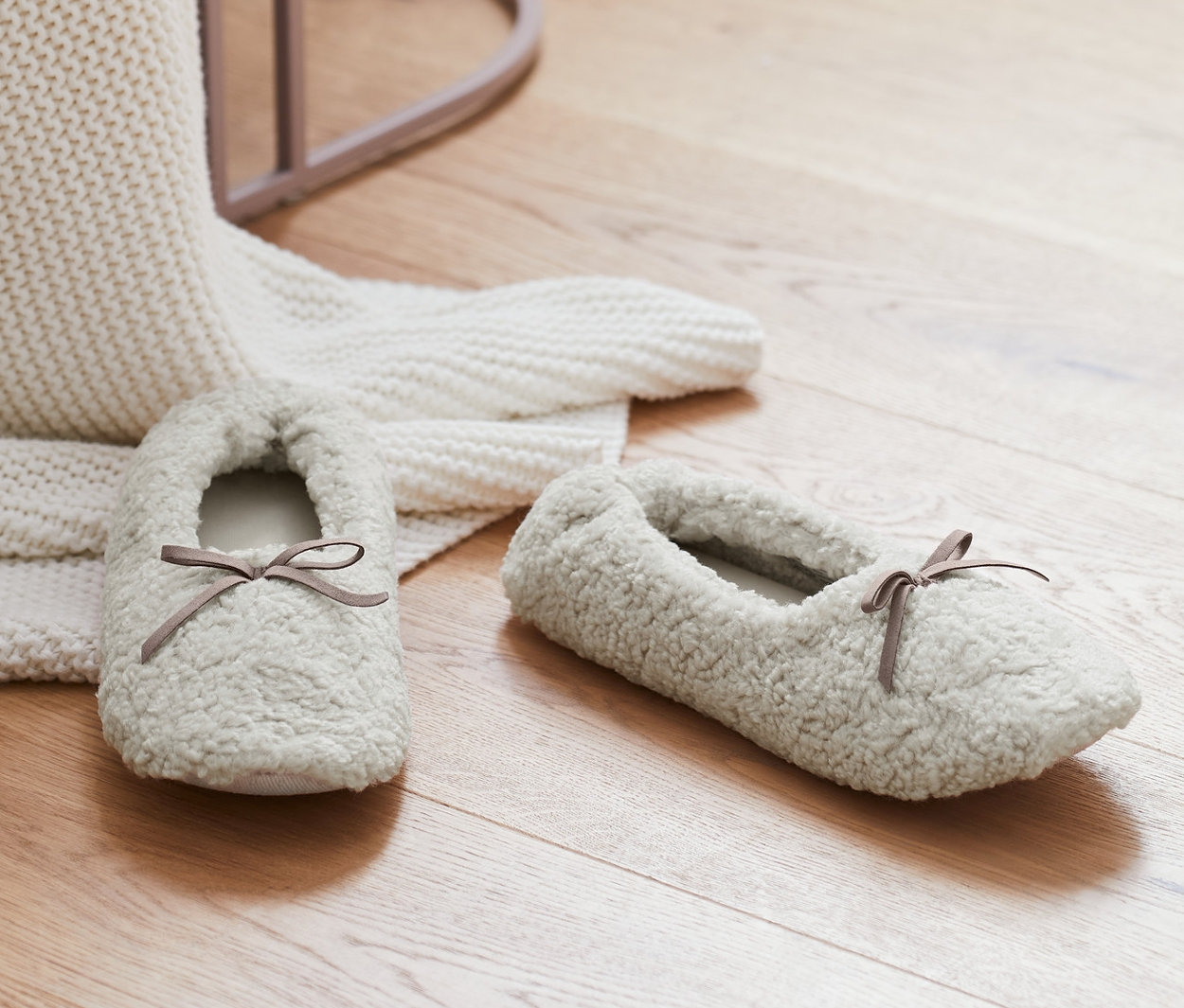 Na drewnianej podłodze leżą kapcie w kolorze kremowym obok białego koca z dzianiny i różowego stołka.