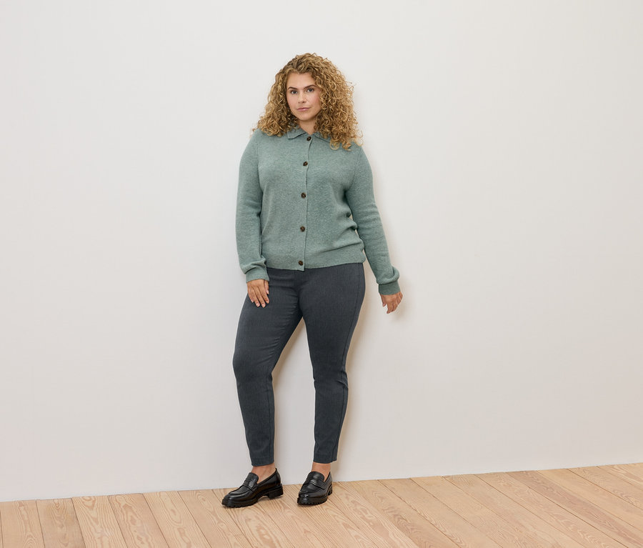Kobieta o kręconych włosach stoi, ubrana w zielony kardigan, szare spodnie i czarne mokasyny.