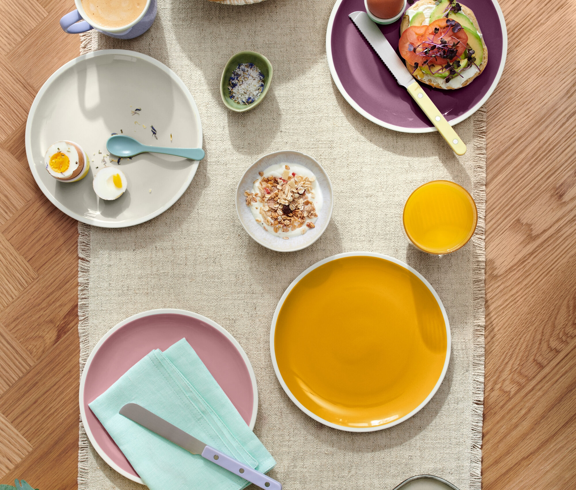 Widok z góry na nakryty stół z różnorodnymi potrawami, w tym pieczywem w koszyku, talerzami z jajkami, tostem z awokado i serem, wraz z napojami takimi jak kawa i sok.