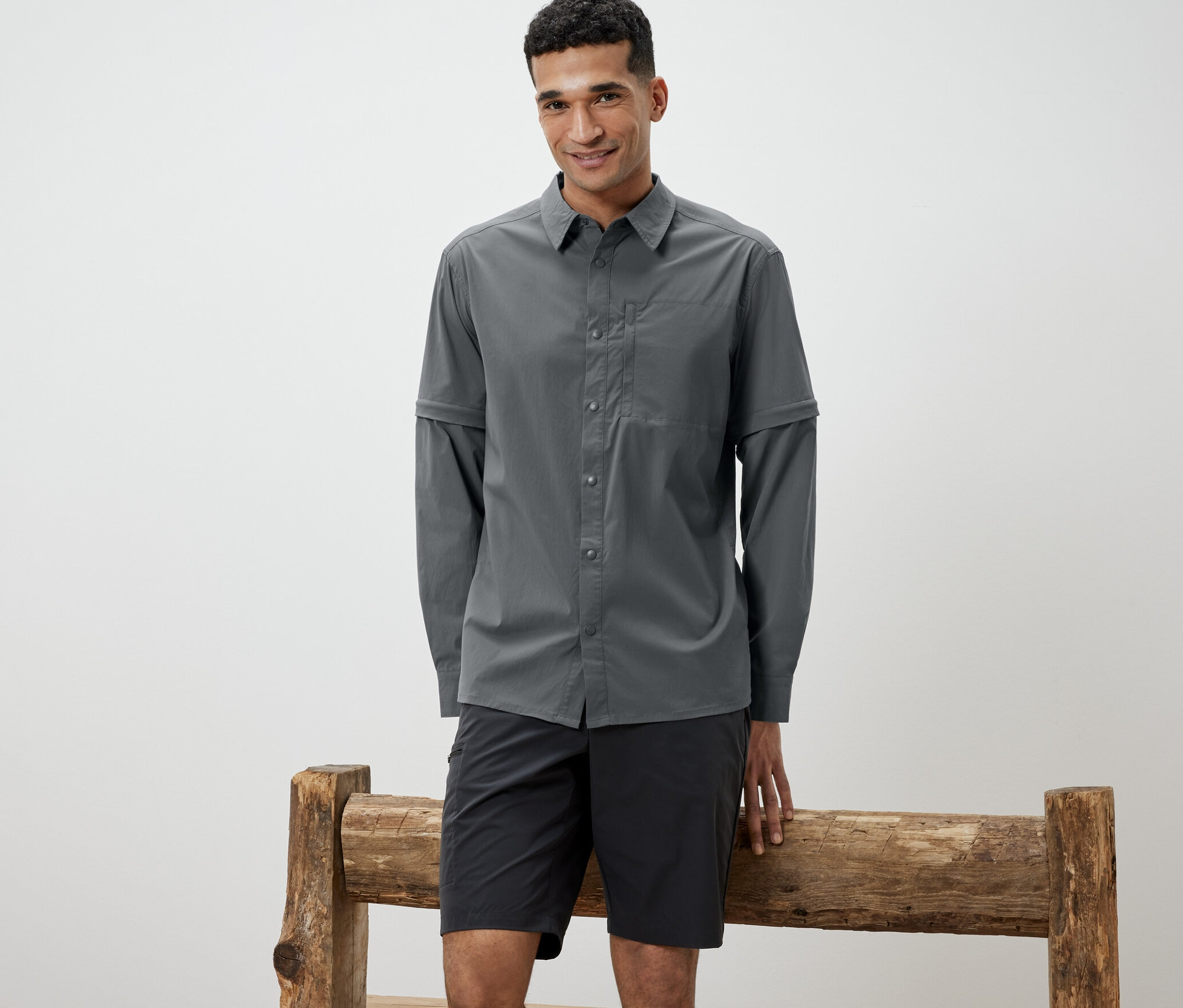 Mężczyzna oparty o drewnianą ławkę ma na sobie szarą koszulę outdoorową i czarne szorty.