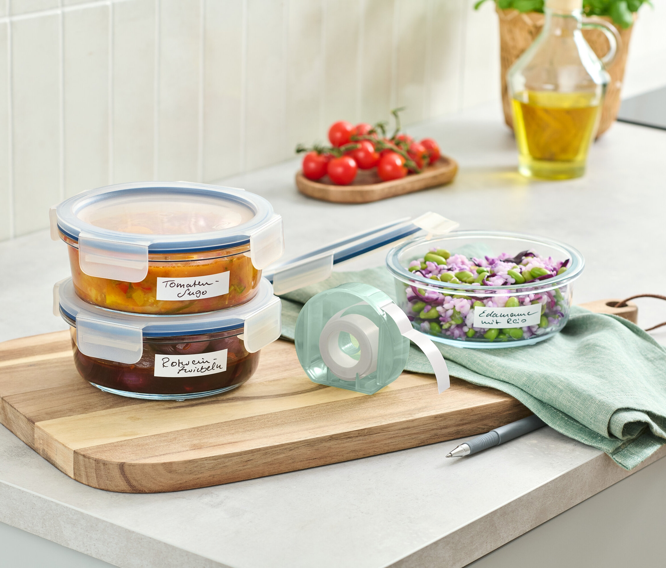 Dwa szklane pojemniki z jedzeniem ułożone na drewnianej desce obok rolki z etykietami.
