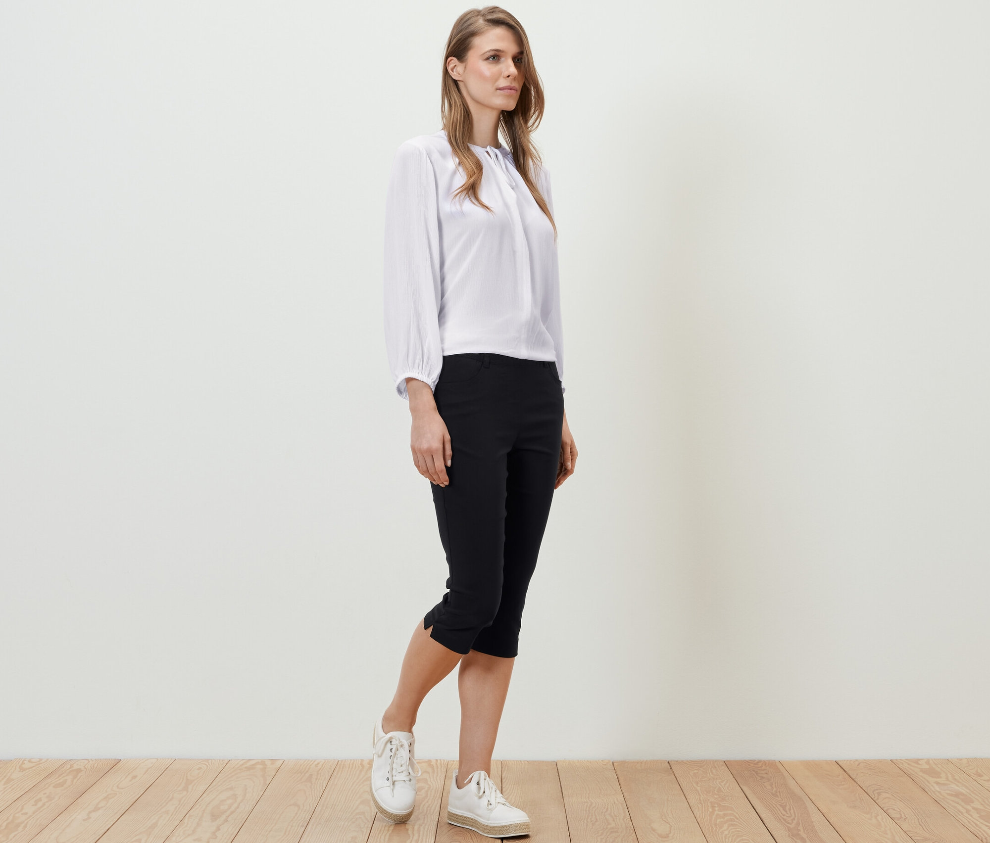 Kobieta stoi na drewnianej podłodze, ubrana w białą bluzkę i czarne spodnie z bengaliny o długości 3/4.