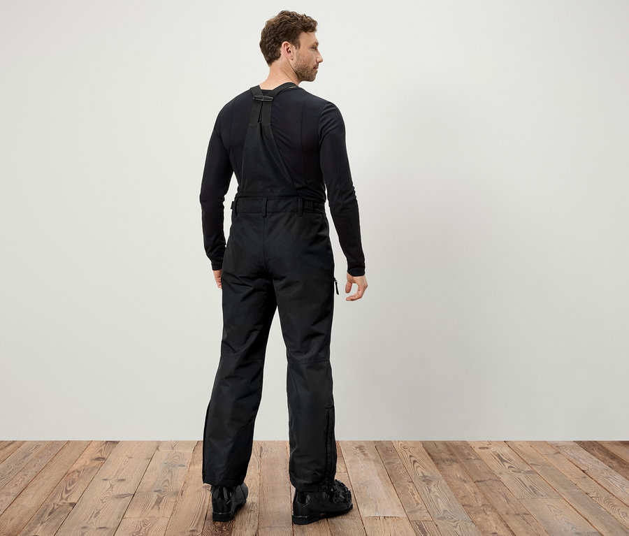 Mężczyzna tyłem do kamery ma na sobie czarne spodnie narciarskie na szelkach i czarne buty narciarskie.