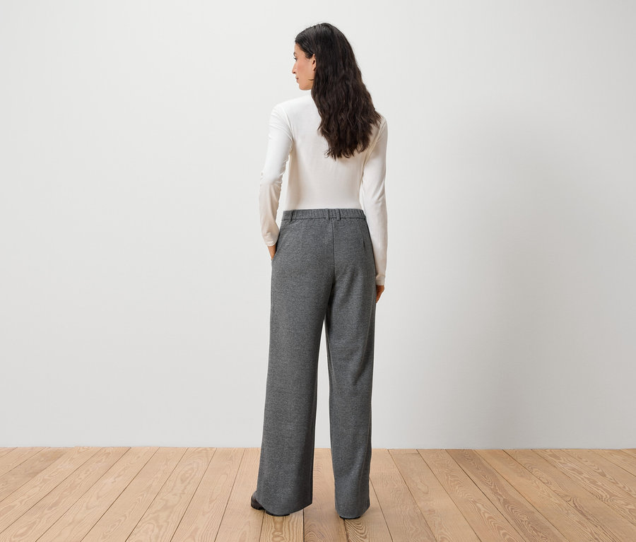 Kobieta tyłem do kamery ma na sobie białą bluzkę z długim rękawem i szare spodnie.