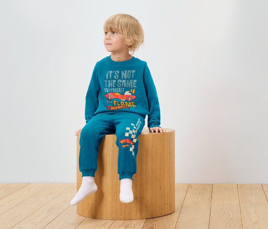Chłopiec siedzi na drewnianym stołku, ubrany w niebieski dres z nadrukiem Hot Wheels.