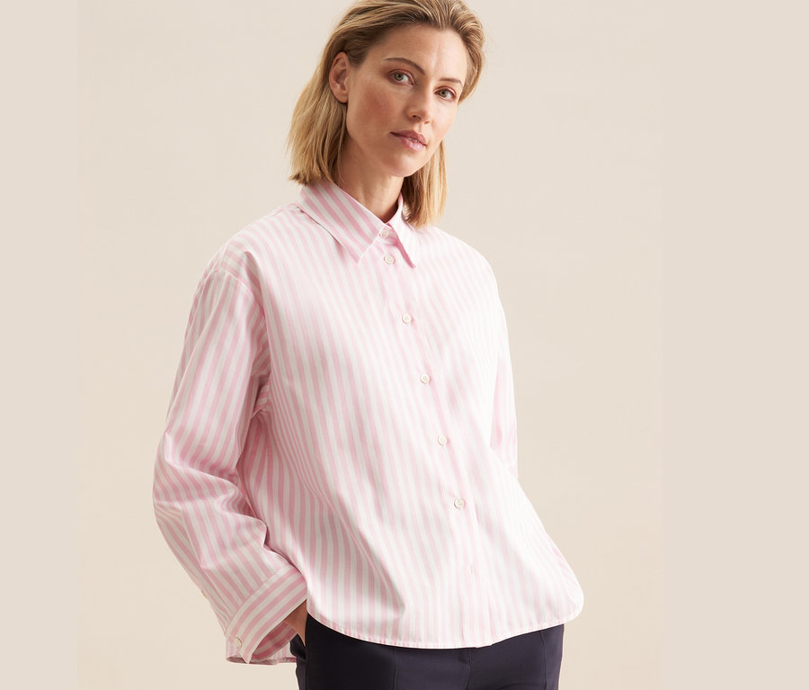 Kobieta ma na sobie różową bluzkę koszulową oversize Seidensticker w paski i granatowe spodnie.