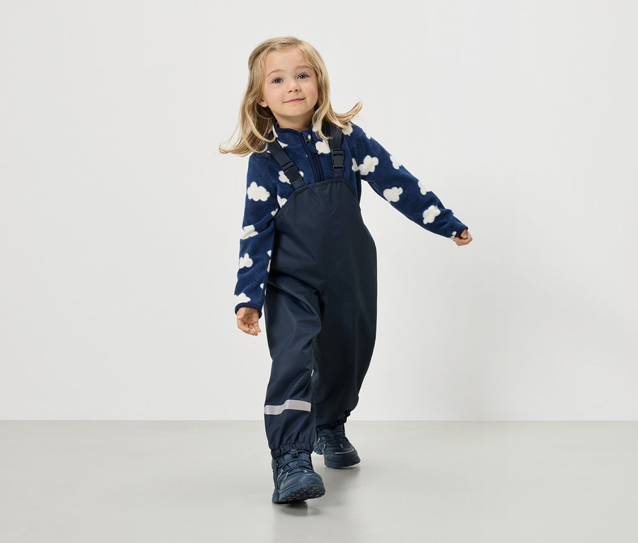 Blondwłosa dziewczynka ma na sobie granatową bluzę polarową w chmury i granatowe spodnie na szelkach.