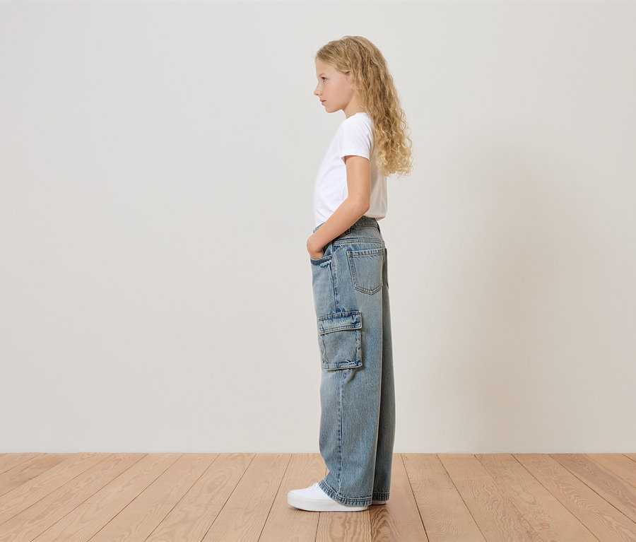 Młoda dziewczyna o blond, kręconych włosach stoi bokiem, ubrana w białą koszulkę, szerokie dżinsy z boczną kieszenią i białe tenisówki.