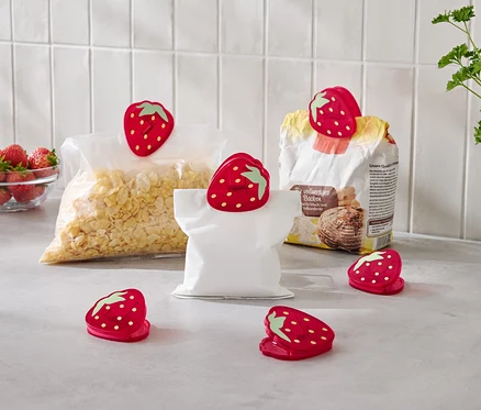 Na blacie kuchennym stoi miska truskawek, torba płatków kukurydzianych, torba mąki i pusta torba, wszystkie zamknięte czerwonymi klipsami w kształcie truskawek.