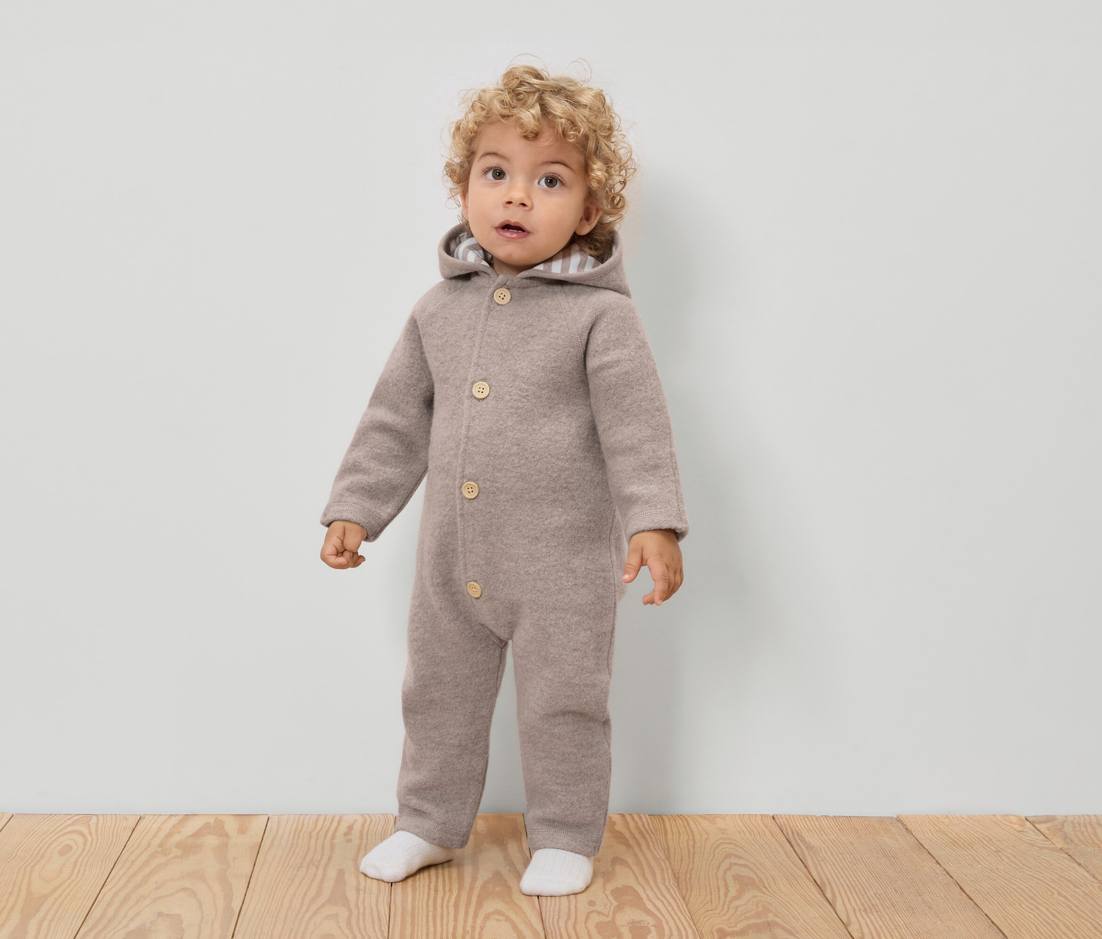 Małe dziecko z kręconymi włosami ma na sobie jasnobrązowy wełniany kombinezon z kapturem i guzikami oraz białe skarpetki.
