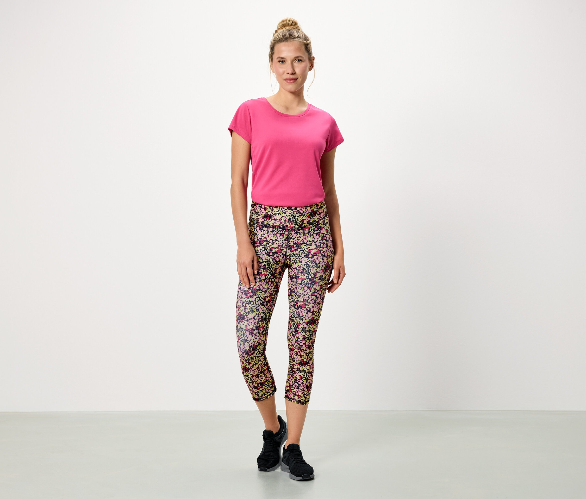 Kobieta stoi na białym tle w różowej koszulce i kwiecistych legginsach.