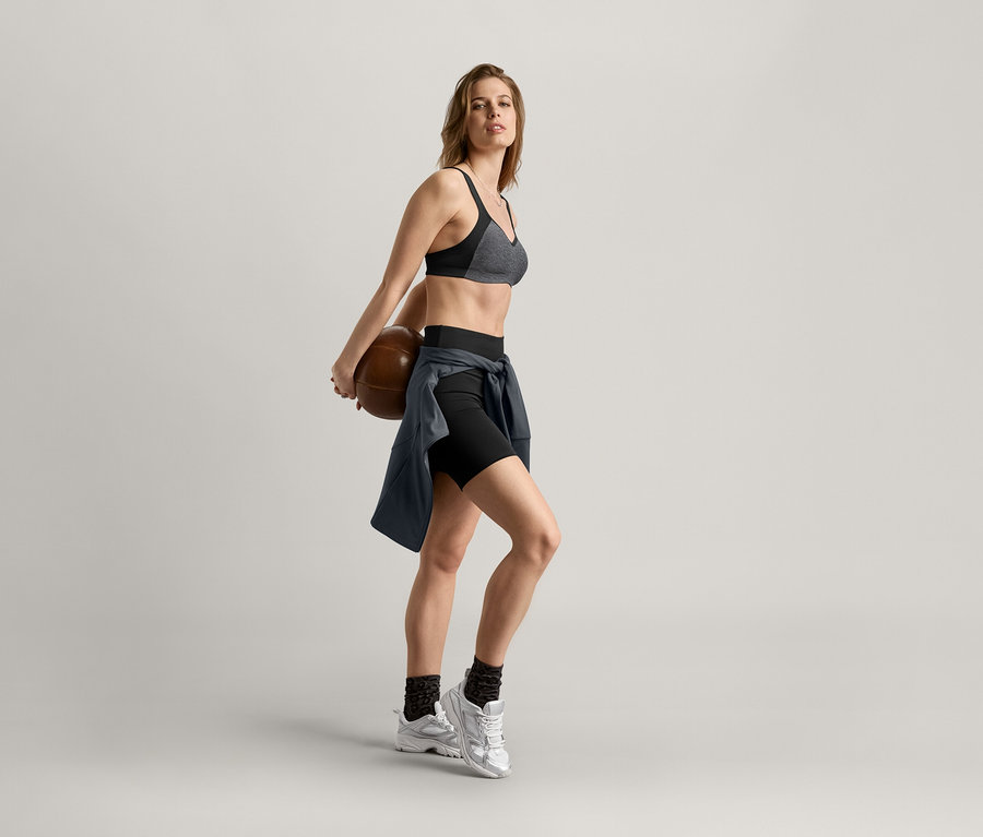 Kobieta trzyma piłkę do koszykówki za plecami, ma na sobie stanik sportowy, szorty i antracytową bluzę sportową.