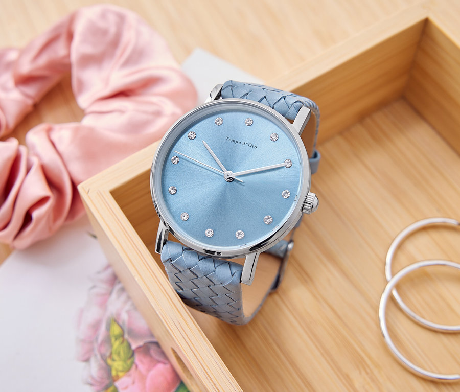 Jasnoniebieski zegarek z dżetami leży w drewnianym pudełku obok różowej gumki do włosów, notatnika i srebrnych kolczyków.