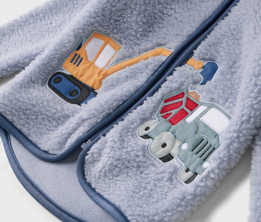 Szczegółowy widok jasnoniebieskiej kurtki polarowej z zamkiem błyskawicznym i naszytymi motywami koparki i ciężarówki.
