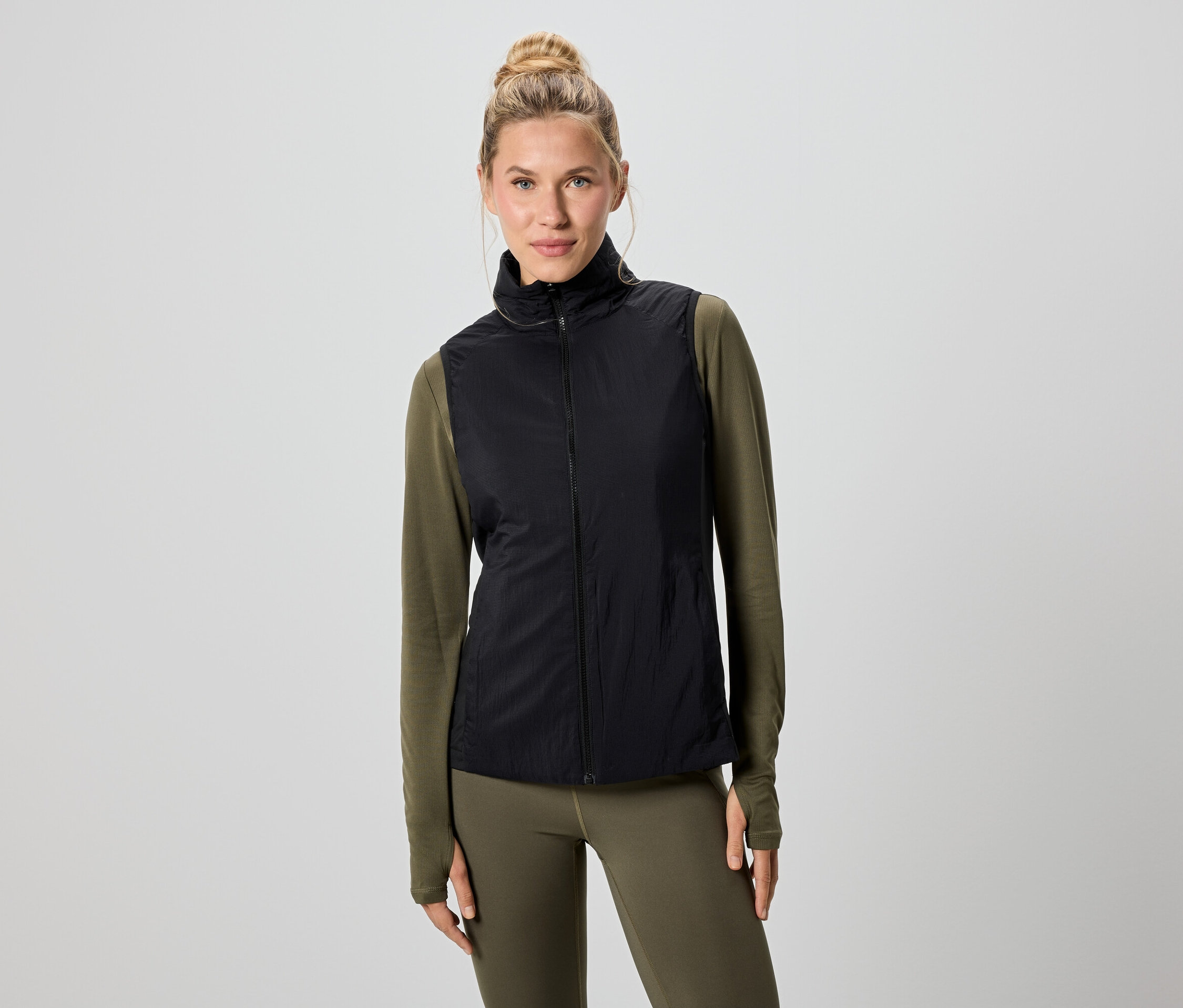 Kobieta o blond włosach ma na sobie czarną kamizelkę nałożoną na zieloną koszulę z długim rękawem i legginsy.