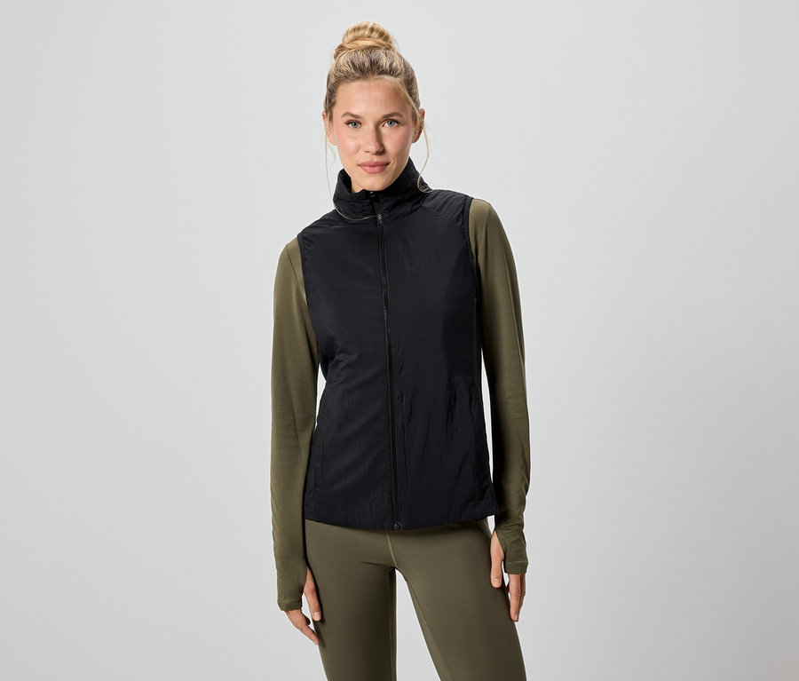 Kobieta o blond włosach ma na sobie czarną kamizelkę nałożoną na zieloną koszulę z długim rękawem i legginsy.