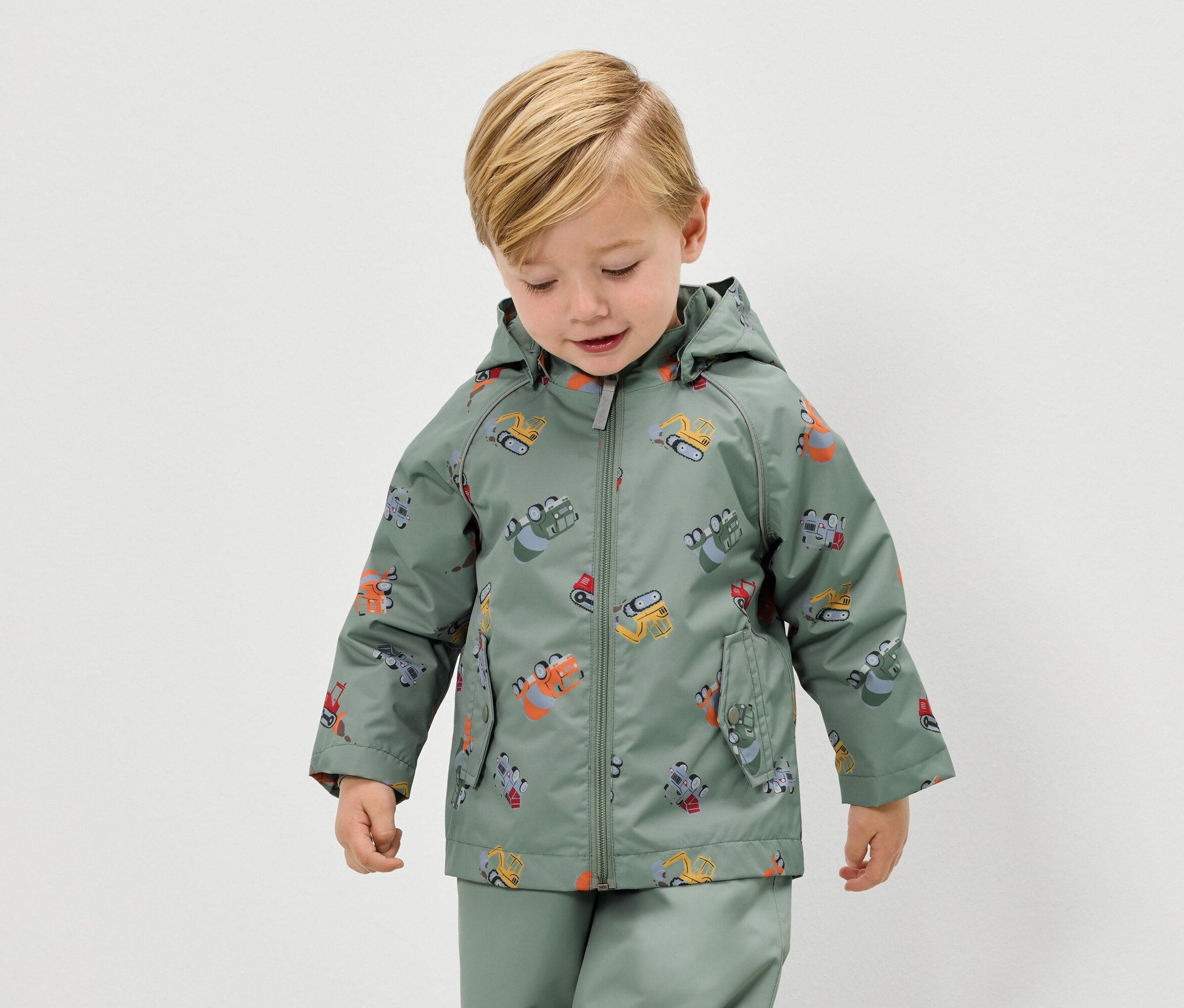 Blondwłosy chłopiec ma na sobie zieloną kurtkę przeciwdeszczową z motywem koparki i pasujące spodnie przeciwdeszczowe.