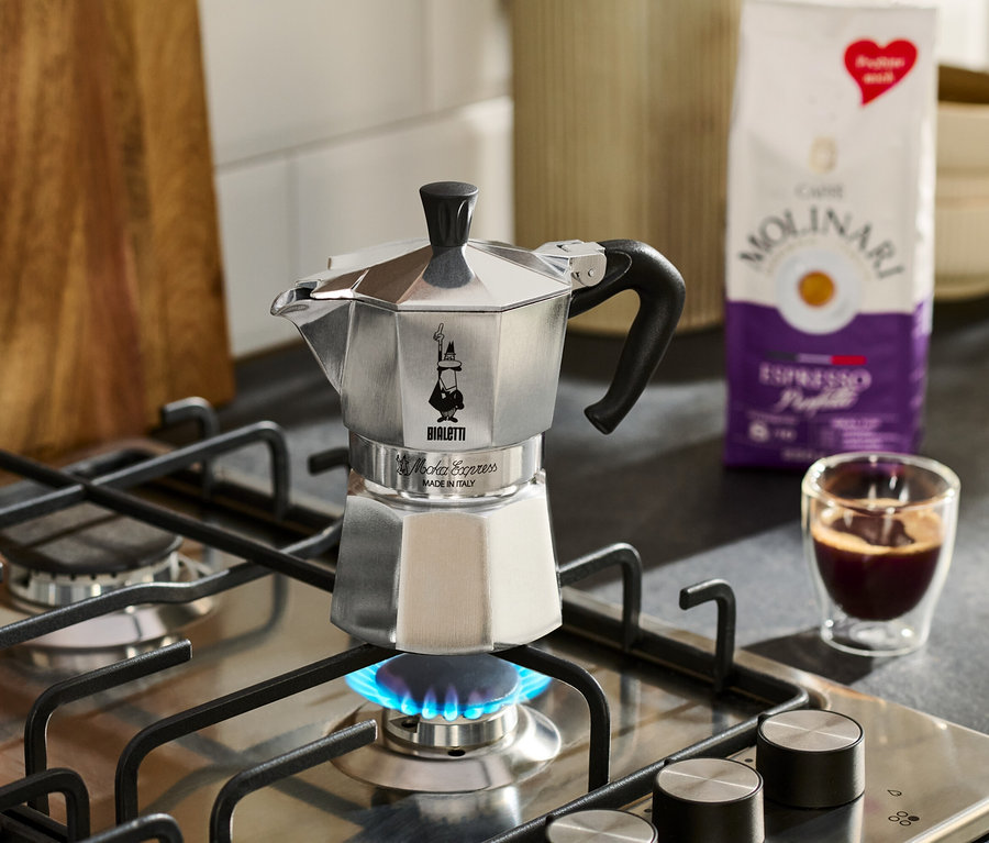 Srebrny ekspres do kawy stoi na kuchence gazowej z niebieskim płomieniem, obok filiżanka espresso i paczka kawy Molinari.