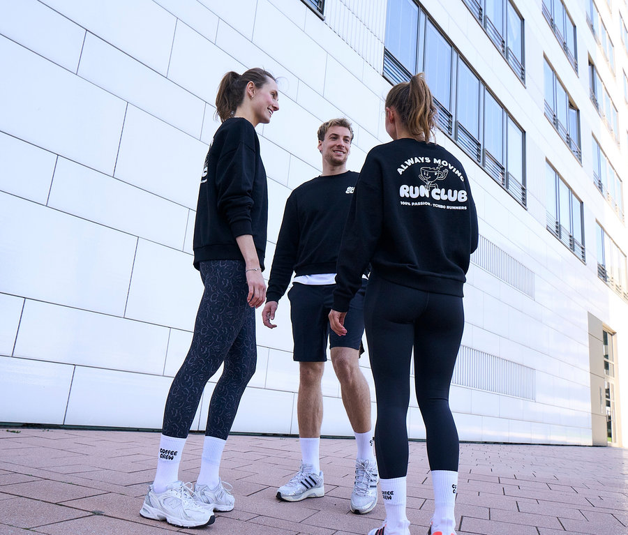 Trzy osoby, dwie kobiety i mężczyzna, stoją uśmiechnięte i rozmawiają. Wszyscy noszą czarne bluzy i białe skarpety tenisowe Coffee Crew unisex. Mężczyzna ma na sobie czarne szorty sportowe. Jedna kobieta nosi szaro-czarne legginsy sportowe „ActivePower”, druga czarne legginsy sportowe „ActivePower”.