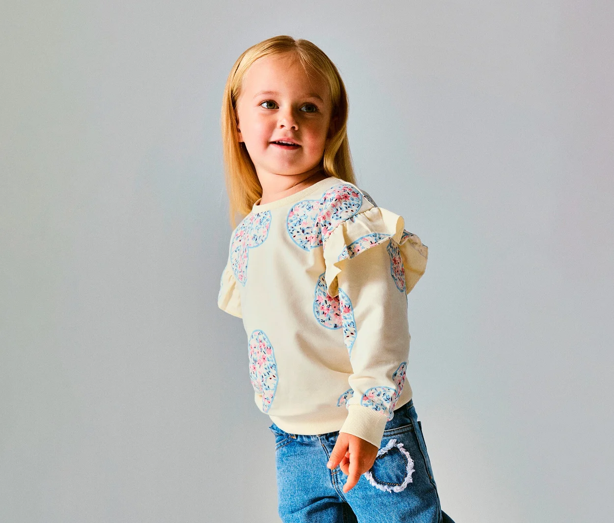 Młoda dziewczynka z blond włosami w kremowym swetrze NAME IT Kinder-Sweater z kwiatowymi wzorami serc i falbankami na ramionach, w niebieskich dżinsach, patrzy w prawo.