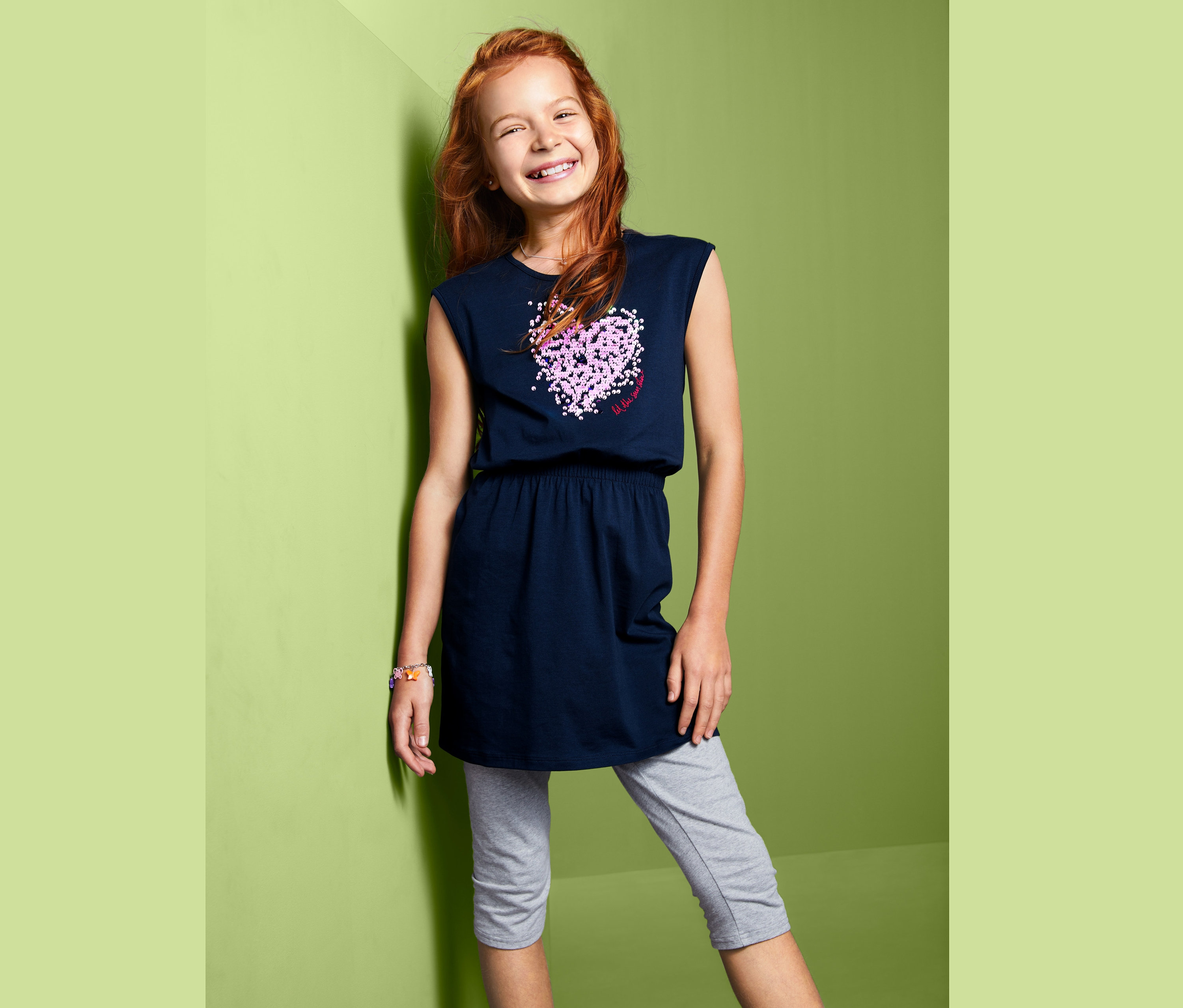 Uśmiechnięta dziewczyna pozuje w granatowej sukience dżersejowej z dwustronnymi cekinami i szarych legginsach.