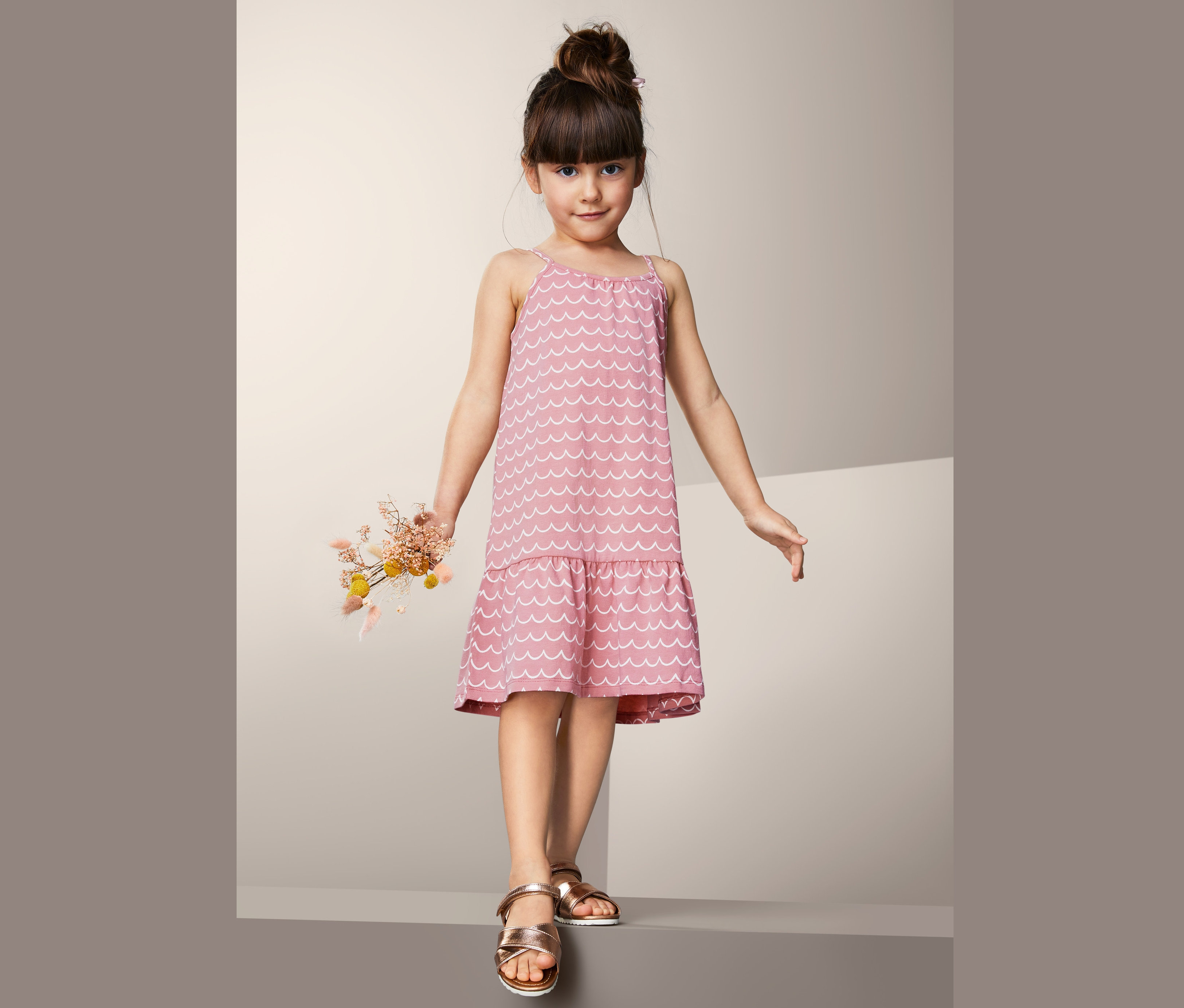 Dziewczynka w różowej sukience z dżerseju z białym falistym wzorem trzyma bukiet suszonych kwiatów.