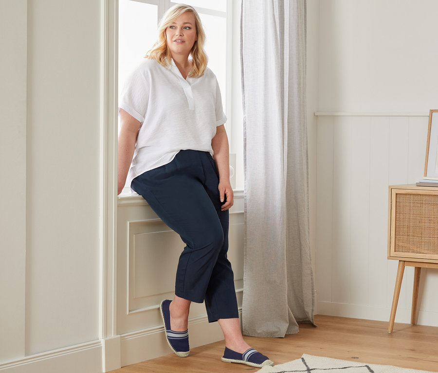 Kobieta siedzi na parapecie w białej bluzce oversized, luźnych spodniach chino i espadrylach.