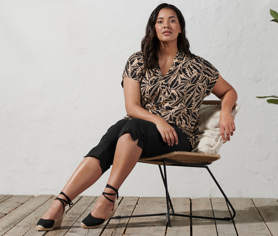 Kobieta siedzi na krześle i ma na sobie espadryle na koturnie, spodnie stretchowe o długości 3/4 oraz bluzkę z nadrukiem i stójką.
