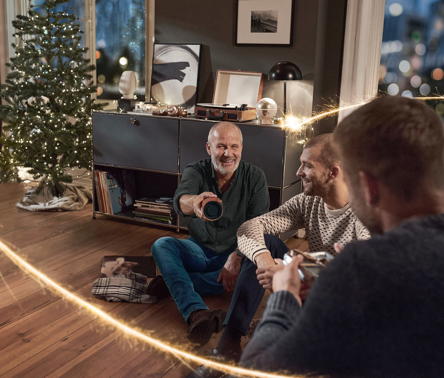 Trzech mężczyzn siedzi na podłodze w salonie, jeden trzyma Głośnik z funkcją Bluetooth® w stylu vintage, z pokrowcem ze sztucznej skóry. Ubrani są w Męskie spodnie dżinsowe, Sweter męski z dzianiny i Koszula męska z lekkiej flaneli.