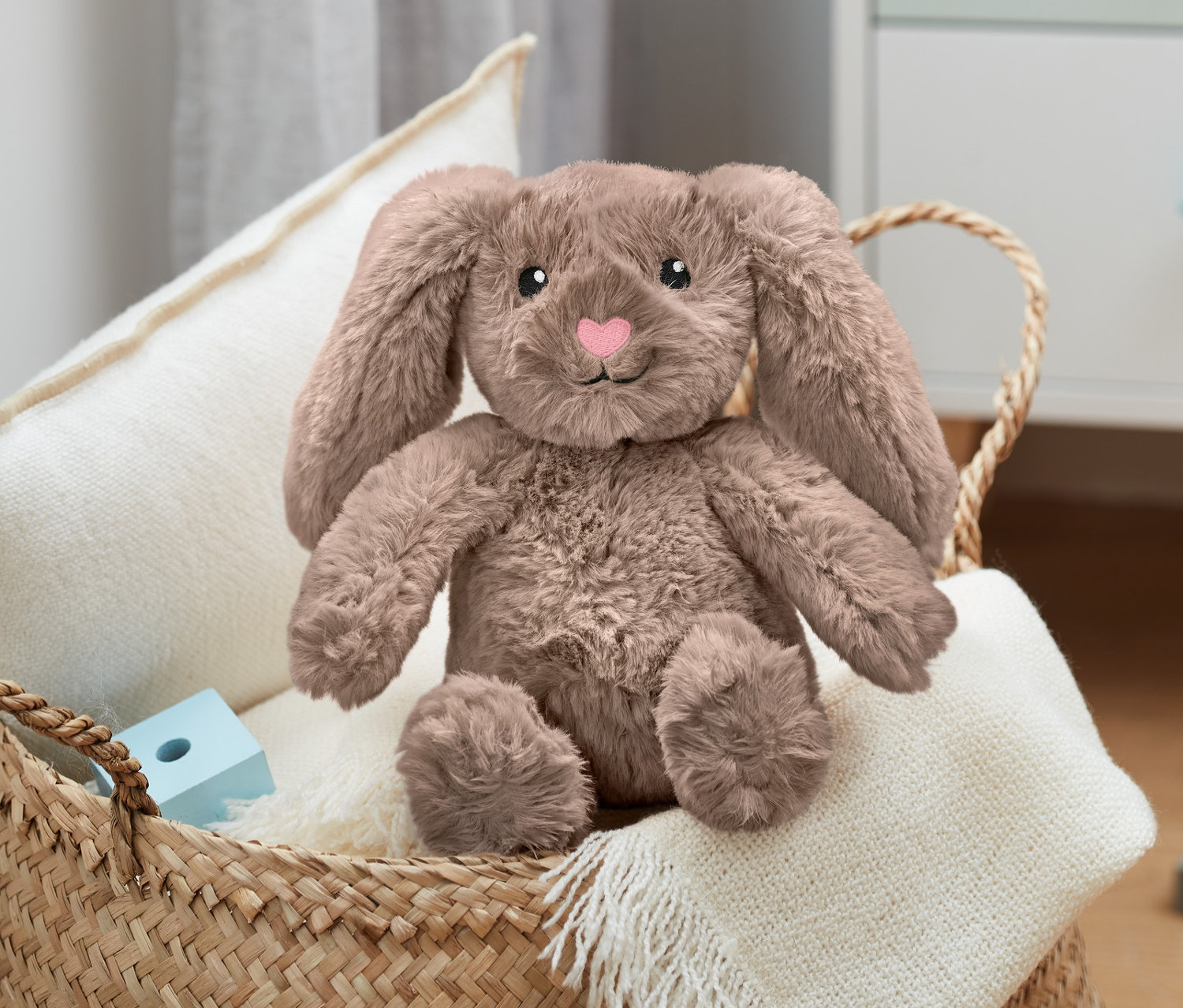 Pluszowy zając siedzi w wiklinowym koszu na białym kocu obok drewnianych klocków.