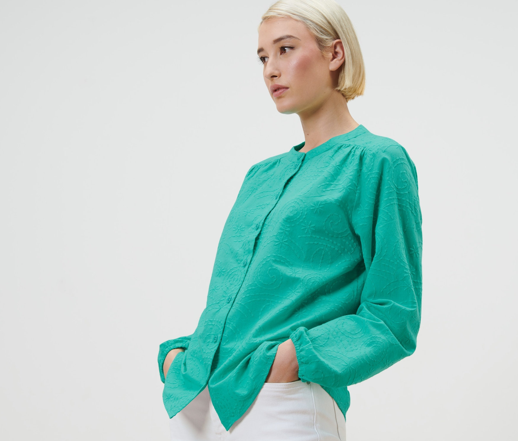 Blondynka pozuje w zielonej bluzce z haftem i białych spodniach.