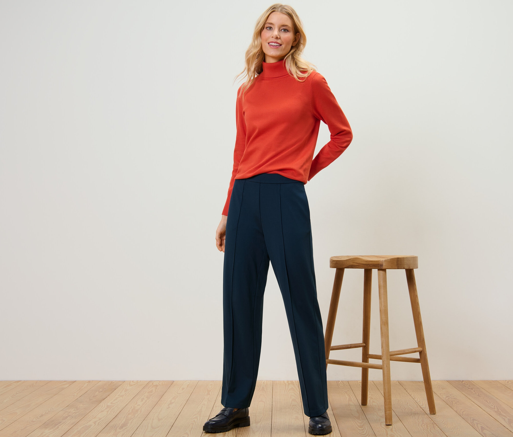 Kobieta pozuje na tle studia, ubrana w pomarańczowy golf i granatowe spodnie dżersejowe z kantem.