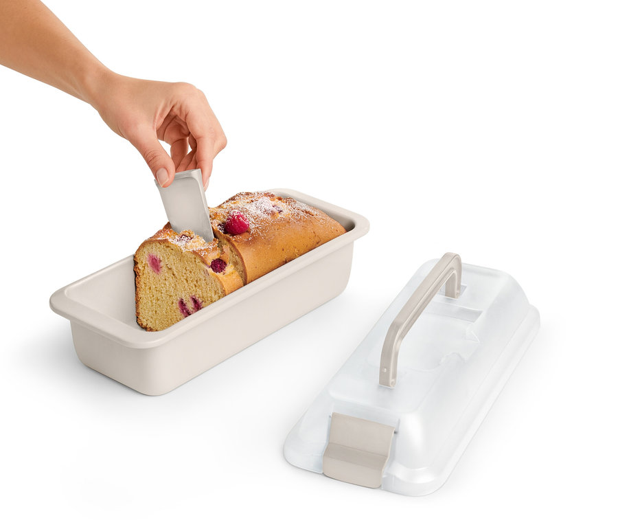 Ręka kroi kawałek ciasta malinowego w prostokątnej formie do pieczenia z pokrywką do transportu.