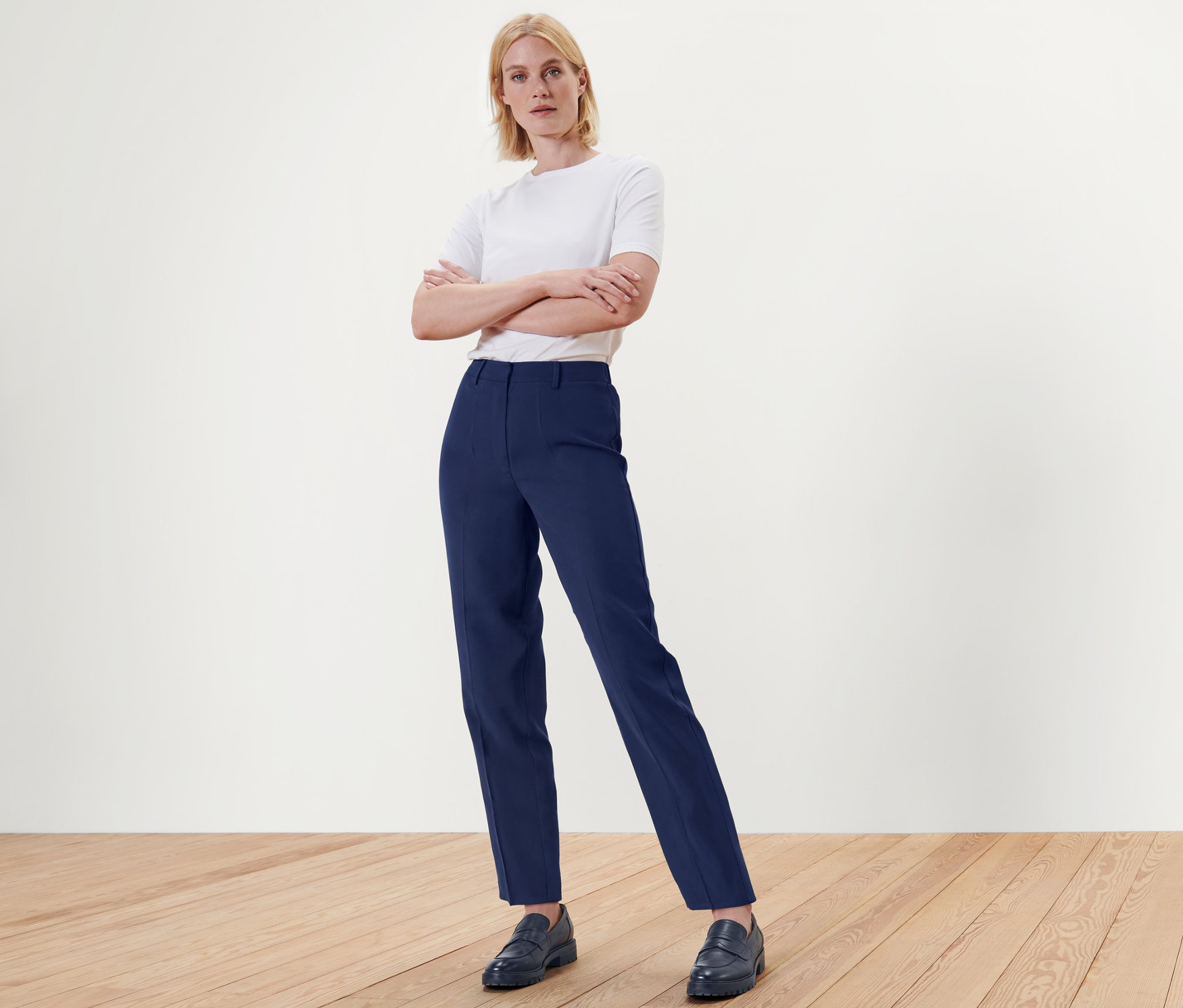 Blondynka stoi ze skrzyżowanymi ramionami w niebieskich spodniach z tkaniny i butach na drewnianej podłodze.