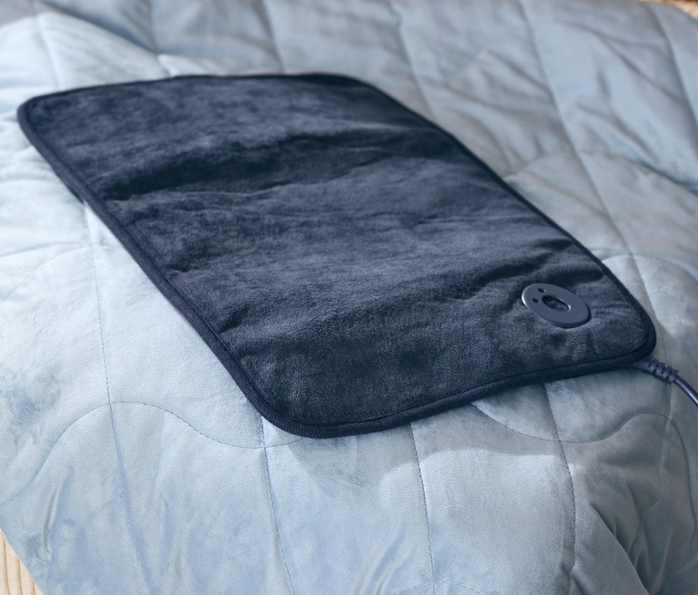 Buty rozgrzewające i mała poduszka elektryczna leżą na odprężającej kołdrze obciążeniowej o wadze 6 kg.